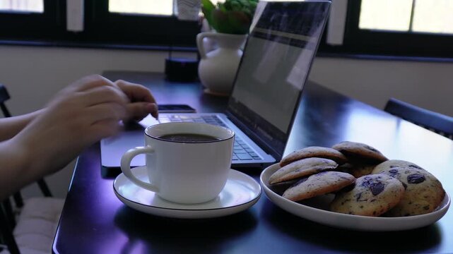 Female hands placing coffee cup on saucer and continuing work on laptop with cookies on desk.