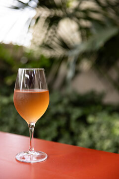 Glass of orange wine on bright red table with lush plants in the background in cafe or patio.