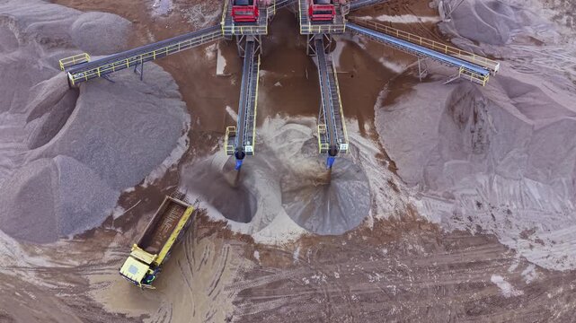 Multiple conveyor belts transfer sand and gravel at a processing site. A yellow truck is nearby, loading materials for transport. Work takes place in an open area during daytime.