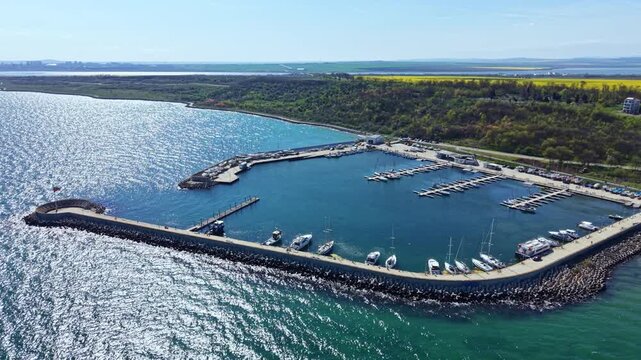 A marina is seen with various boats and yachts docked. The water is clear and reflects sunlight while land with trees borders the shore. Above, the sky is bright and blue.