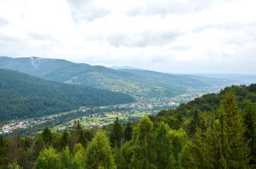 Fototapeta premium Panoramic green mountain valley with Yaremche village nestled among dense forest and rolling hills, showing rural landscape, misty atmosphere and cloudy sky. Carpathian Mountains, Ukraine