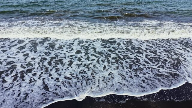 Waves flow onto a dark sandy beach during the day. The ocean is blue and the foam from the water creates patterns as it recedes, bringing life to the shore.