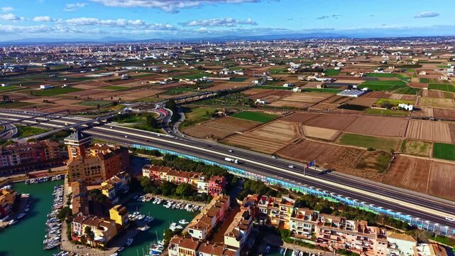 Fields stretch across the land under a blue sky. A road runs through the scenery with few cars visible. Buildings sit close to a water body, showing human activity in the area.