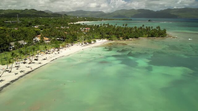 Aerial panorama of tropical bay in Las Galeras with palm trees, green hills, clear blue water and mountains in the background filmed in bright daylight.