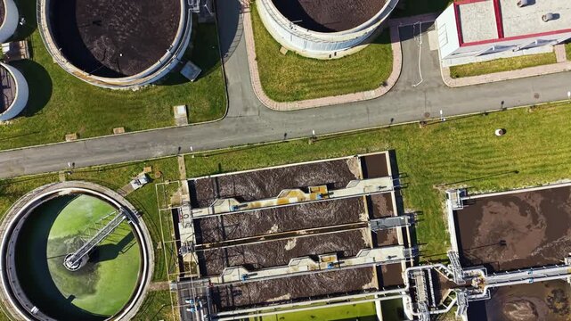 Workers and machines process wastewater at a treatment plant. Large circular and rectangular tanks are visible. The area has roads, grass, and different sections for treatment processes.