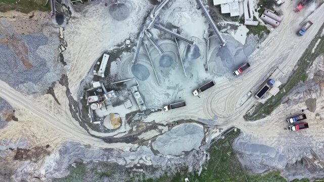 Trucks move around a granite quarry site where machinery processes stone and material. Workers manage equipment and transport loads. Dust rises from the ground.