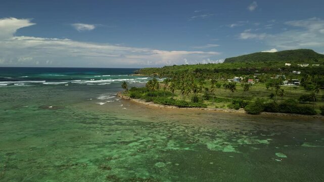 Aerial panoramic view of Las Galeras coastline with lush tropical greenery, distant mountains, palm trees and clear turquoise sea under bright sunny skies in 4K drone footage