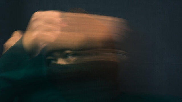 A male person wearing a dark brown balaclava faces forward while a sudden hand movement creates thick horizontal blur sweeping across the frame. The disorienting visual effect conceals