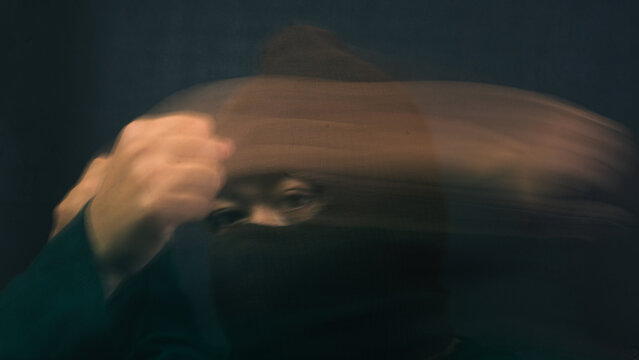 A mysterious male figure wearing a brown balaclava is heavily obscured by extreme horizontal motion blur from his hand sweeping across his face. The aggressive and fast movement creates