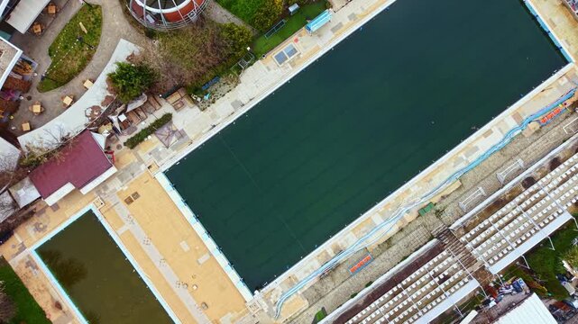 A large swimming pool area is visible from above, surrounded by green spaces and seating. Two pools are in the scene, with people relaxing and a bird flying over the water.