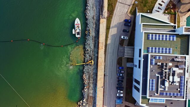 A boat is seen near the shore where construction equipment is working. People are near a modern building with solar panels, parked cars visible on the street.