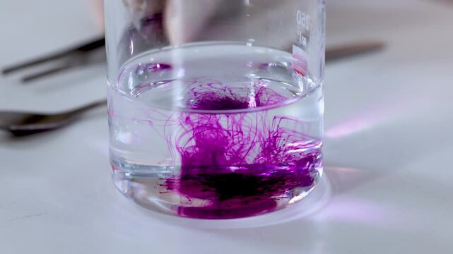 A close up view of potassium permanganate dissolving in water in a beaker, forming a vibrant purple solution while being stirred with a spoon.