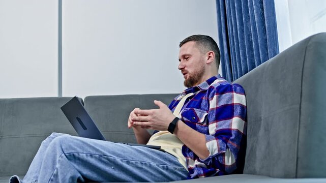 A man sits on a couch in his living room while using a laptop. He gestures as he speaks during a virtual meeting in the afternoon sunlight coming through the window.