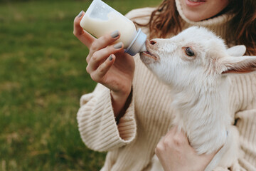 Bright outdoor moment with a woman bottle feeding a curious goat kid, warm light, cozy sweater, relaxed posture, lush grass background, gentle bond, natural colors, simple rural scene, wholesome © SHOTPRIME STUDIO