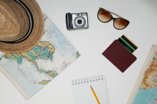 Flat lay of travel planning essentials including map, camera, sunglasses, hat, wallet and notebook on a clean white background with copy space.