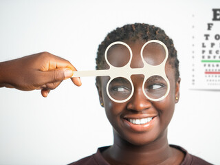 A smiling African woman receives a vision test with an optical flipper lens. A Snellen eye chart hangs in the background of this bright, professional medical clinic setting. © RODWORKS