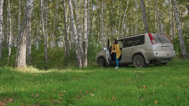 Camper unloading gear from suv trunk, female in yellow jacket lifting cooler and crates, organizing supplies on grass beside birch trees, practical prep for campsite setup, focused movement