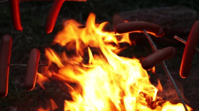 Roasting tasty sausages over bonfire in darkness outdoors, closeup. Camera moving left