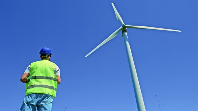 A windmill engineer looks up at a large wind turbine located in a rural area. The engineer wears a safety vest and a helmet while checking the turbine during daytime.