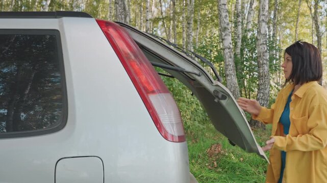 White woman opening car trunk camping in birch forest, lifting folding table into rear cargo, wearing yellow jacket, sunlight filtering through leaves, calm unpacking and setup scene