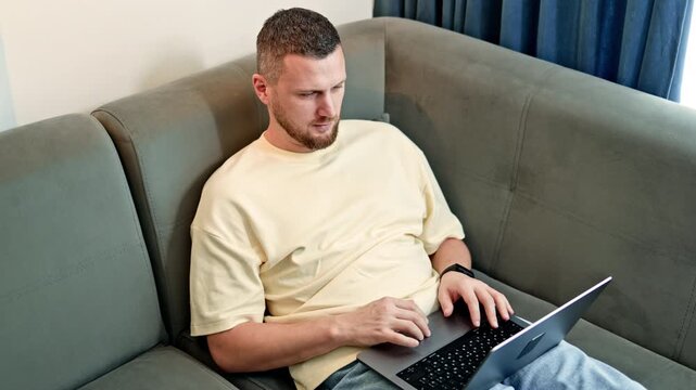 A man sits on a gray couch with a laptop. He types on the keyboard while focusing on the screen. Sunlight enters through a window, illuminating the room.