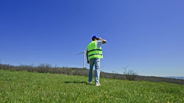 A windmill engineer walks on the green grass of a field while holding a blue helmet. Wind turbines are visible in the distance against the clear sky at midday.