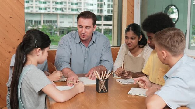 Smart teacher talking and explaining work while multicultural children drawing or taking a note in paper at table with pencil. Happy diverse student drawing or sharing idea while sitting. Edification