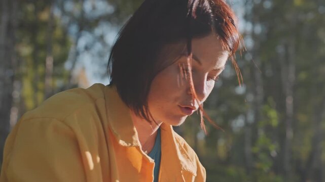 Woman in yellow jacket camping in birch forest soft backlight on short hair closeup candid portrait thoughtful expression warm afternoon sunlight filtering through leaves peaceful outdoor mood