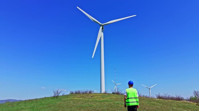 An engineer inspects windmills in a field. The engineer walks on green grass near a wind turbine. The sky is clear and blue. The activity is during the daytime.