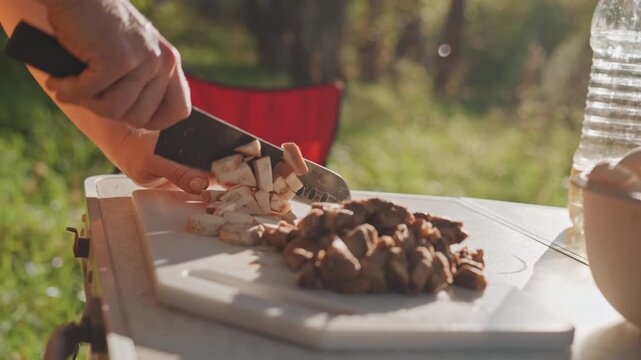 Caucasian hands chopping bread cubes outdoors on plastic board, warm backlight and portable stove nearby, pile of cubed bread and cooked meat for stuffing or croutons, cozy group meal atmosphere