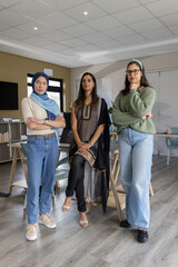 Fototapeta premium Diverse female coworkers standing in meeting room wearing hijab and tunic, holding laptop
