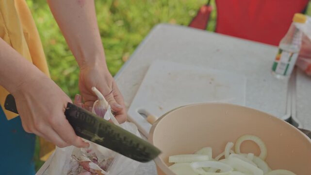 Woman shaving onion into bowl camping table outdoors, closeup on peeler and knife, steady hands forming neat rings, soft sunlight and green backdrop, focused rhythm for camp stew preparation,