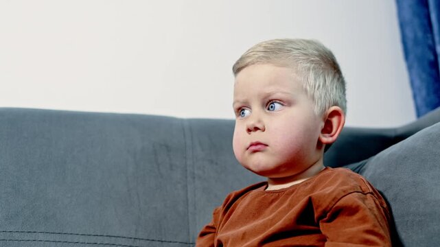 A young child sits on a gray couch with a thoughtful look, gazing forward while surrounded by a simple room filled with soft light. It is a common moment in daily life at home.