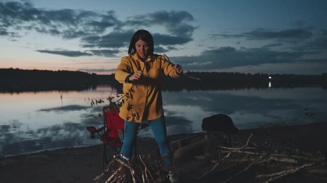 Dusk lakeside camping woman lighting campfire with match and tinder beside calm reflective lake, branches and folding chair nearby, sparks and warm glow contrast cool evening, quiet joyful moment