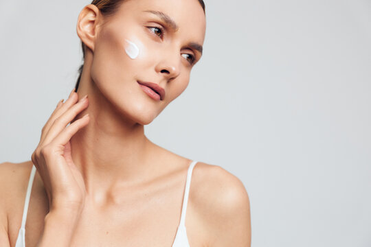 Young woman with clear skin applying white face cream on cheek, wearing white tank top, looking away on light gray background, concept of skincare, beauty, and self-care routine.