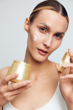 Woman applying white face cream, holding a jar and tube, closeup portrait on light background, beauty skincare routine, wellness and self care, soft studio lighting