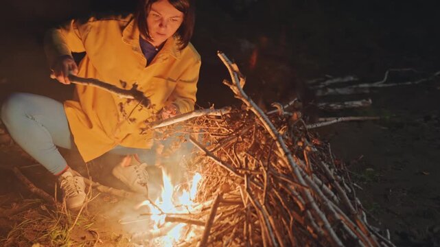 Night camping beach camper stoking fire with long stick, adding driftwood and tending embers, warm glow on yellow jacket, steady motion and focused outdoor ritual, practical camping skill
