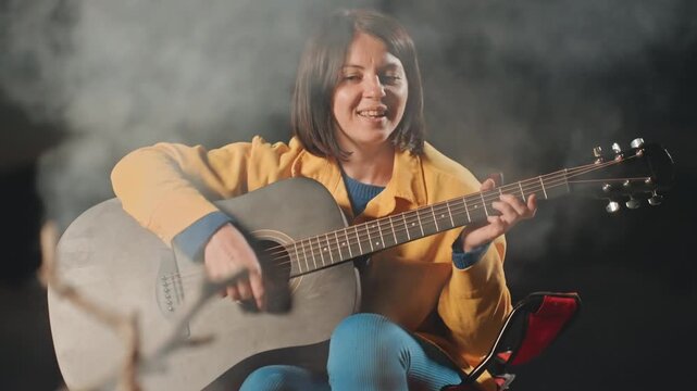 Cozy musical evening. Relaxed lady tunes guitar at sunset. Serene scene of woman singing near lasting fire. Intimate moment with woman serenading beside glowing campfire in peaceful night