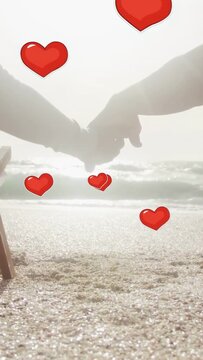 Vertical video: Senior couple reaching hands on sand, causing red hearts floating, promoting health