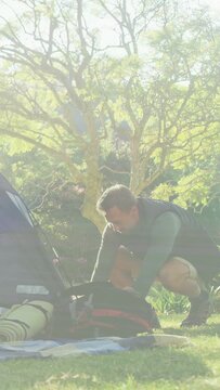 Vertical video: Mature man crouching, unzipping backpack beside tent, unpacking gear for camping