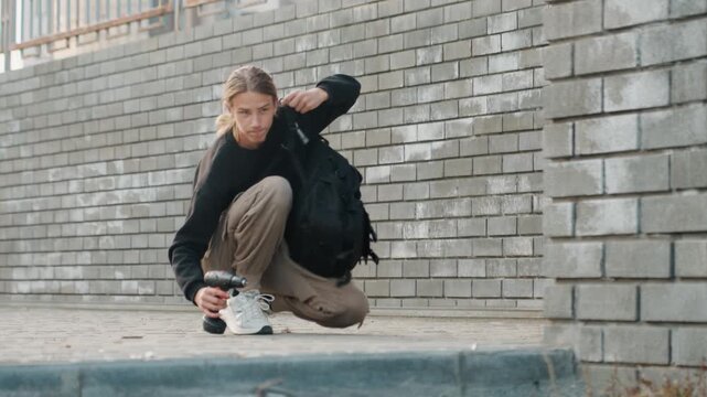 White man crouching by brick wall checking backpack, inspecting zipper near sneakers, cargo pants and black sweater, urban sidewalk, soft overcast light, introspective student vibe, moment of pause