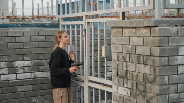Young blonde inspecting metal gate at brick wall, tentative approach and close evaluation of lock and bars, handheld drill at side, calm overcast light, attentive maintenance vibe with subtle urban