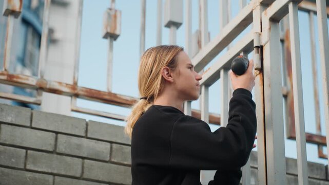 Young blonde securing gate with drill at brick perimeter, decisive upward reach, tightening fasteners and reinforcing bars, handson craft and steady concentration under open sky, practical urban