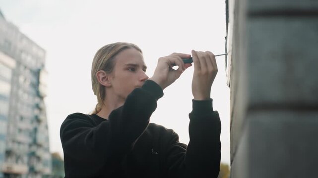White man inspecting small lock on bridge wall, urban waterfront and modern building backdrop, focused hands using screwdriver, methodical motion and precise adjustment in daylight
