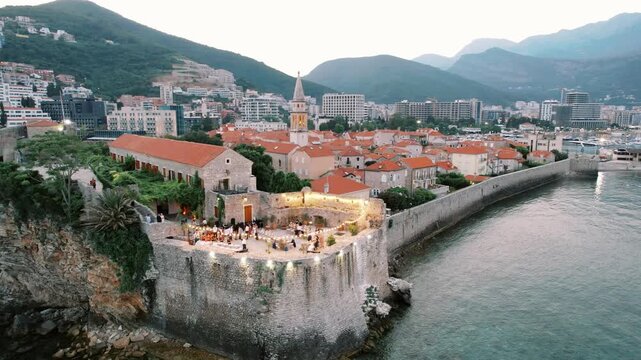 Nightlit fortress overlooking sea and mountains. Luminous fortress on cliffs with diners and scenic silhouettes