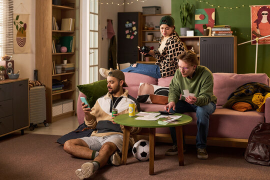 Wide shot of multiethnic students checking smartphone, reviewing papers, managing shared expenses in apartment. Useful for budgeting, student lifestyle, roommate finance marketing