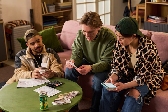Young adult students reviewing bills, counting cash, checking smartphone, calculating shared expenses at home. Ideal for budgeting, student finance, rent splitting, cost of living themes