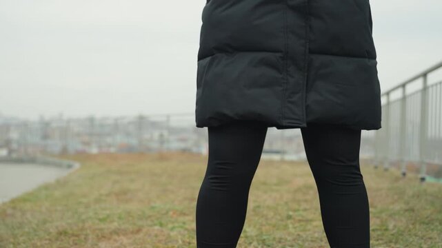 South asian woman close up legs dancing on grass, black leggings and boots, puffer coat hem visible, steady rhythmic steps near railing and waterfront, damp grass texture, focused tempo and balance