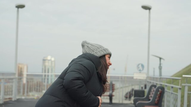 Woman beanie rooftop practicing dance moves, commuter warmup with subtle footwork and shoulder rolls, winter puffer jacket, cloudy city skyline, bench and railing, quiet reflective energy