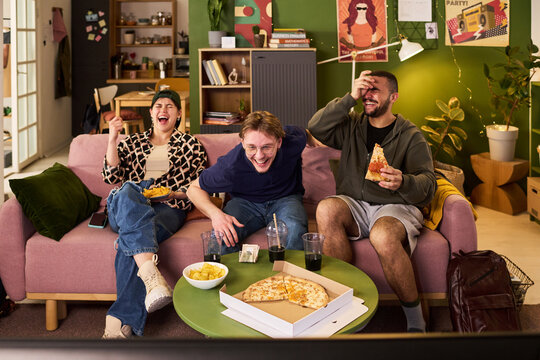 Multiethnic college students laughing while watching television, eating pizza in shared apartment, expressing friendship and leisure. Ideal for youth lifestyle or student housing marketing